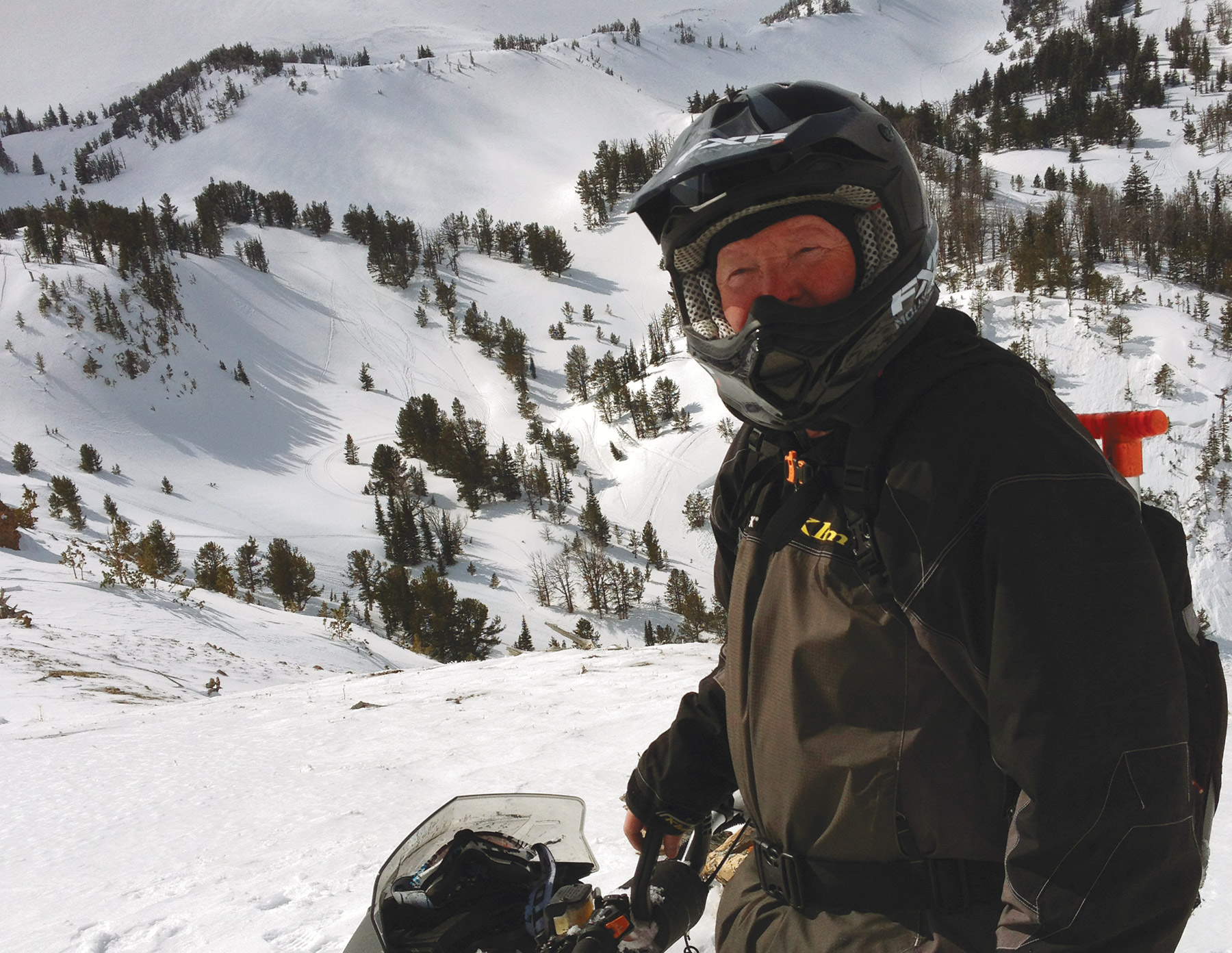 Steve Janes wearing a helmet and ski gear on a snowy ski slope with a snowmobile
