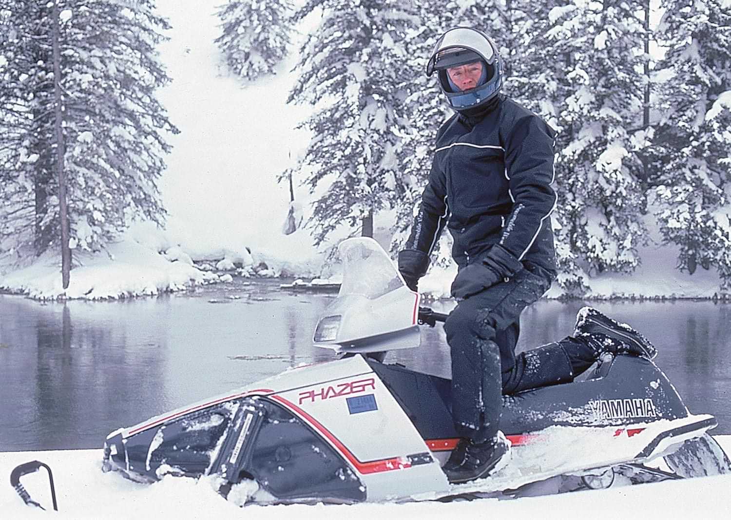 Darryl Harris, wearing a snow suit and helmet with a lifted goggle  is pictured on a Yamaha Phazer parked beside a lake surrounded by snow covered pines