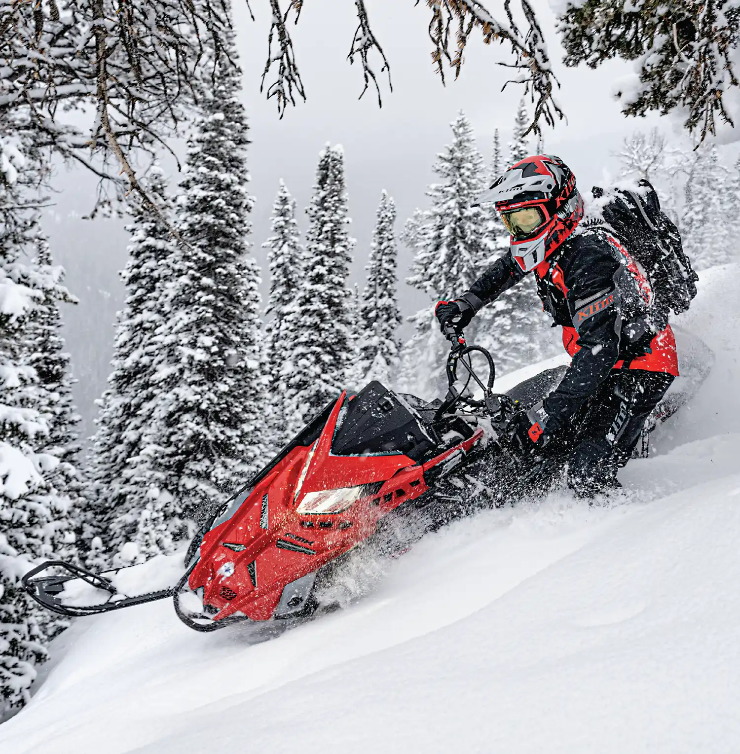 rider taking red snowmobile through trees while wearing a matching red and black snowsuit
